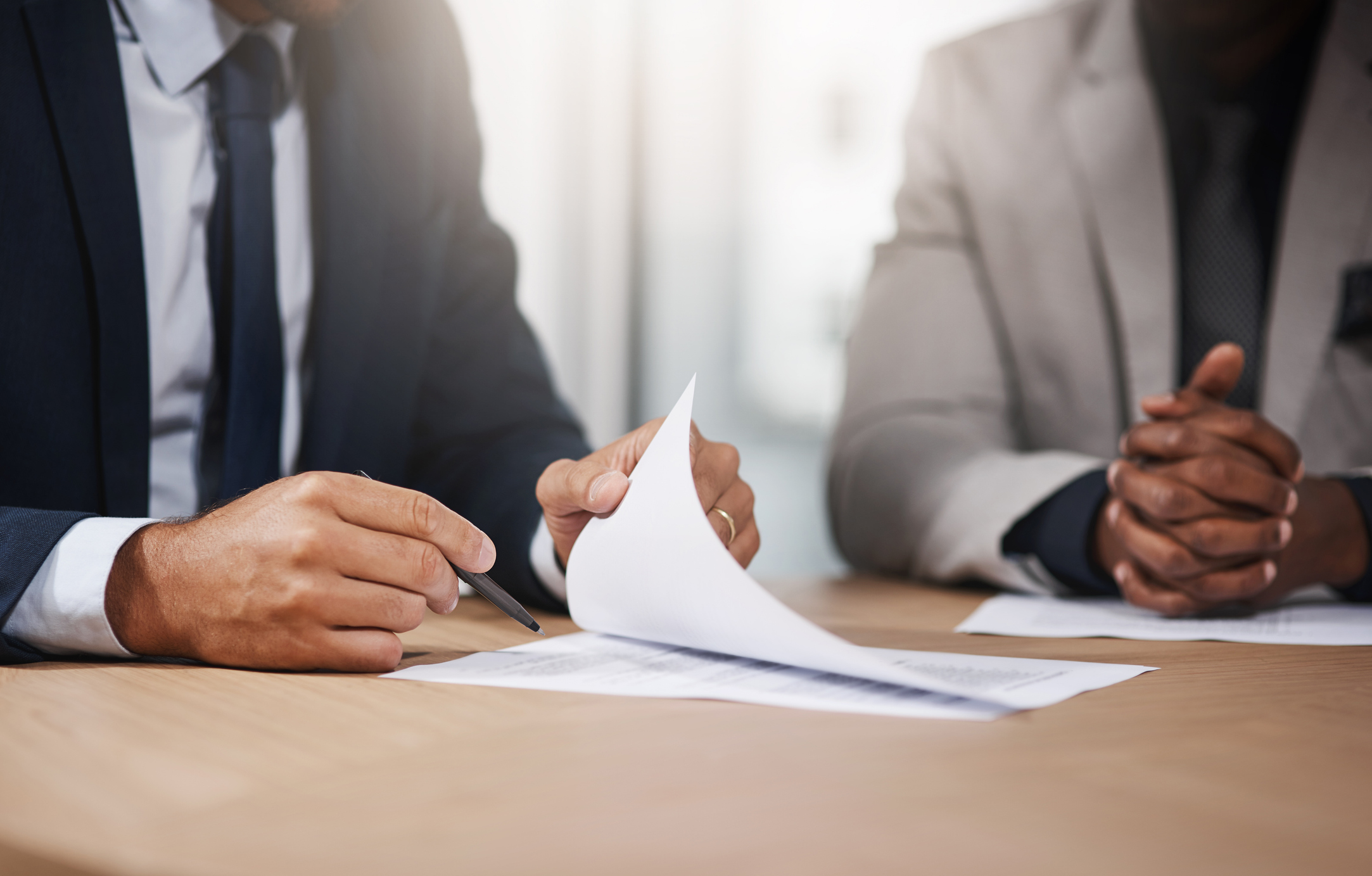 Zwei Personen in Businesskleidung sitzen an einem Tisch, eine Person blättert und unterschreibt ein Dokument.